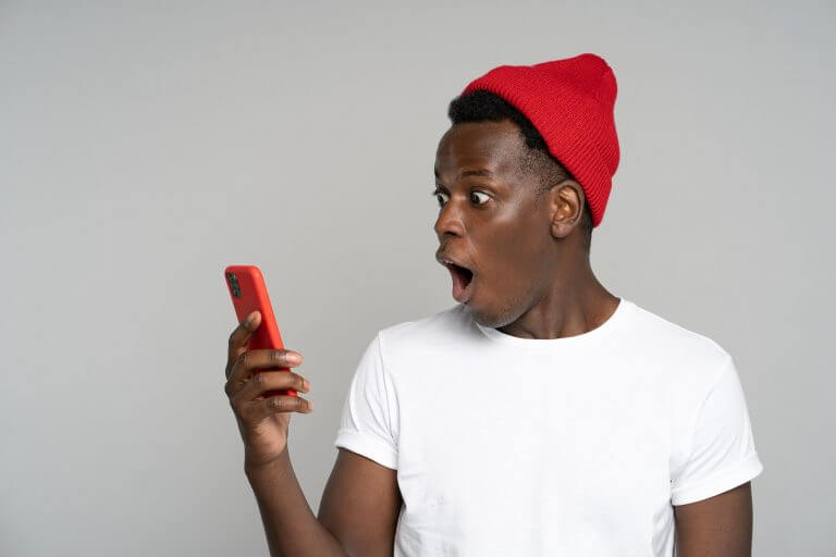 Surprised afro man makes big eyes opens mouth widely, shocked with indecent photo, looking at mobile